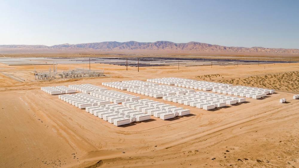 Aerial view of rows of Tesla Megapack battery units at a utility-scale energy storage site in the desert, with a solar array in the background