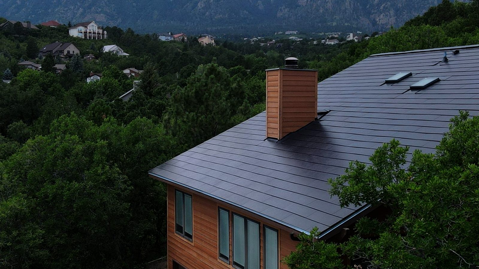 Tesla Solar Roof tiles on a two-story home surrounded by trees, with a mountain backdrop