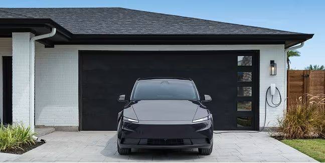 Tesla Wall Connector mounted beside a garage on a home exterior, with a Tesla vehicle parked in the driveway